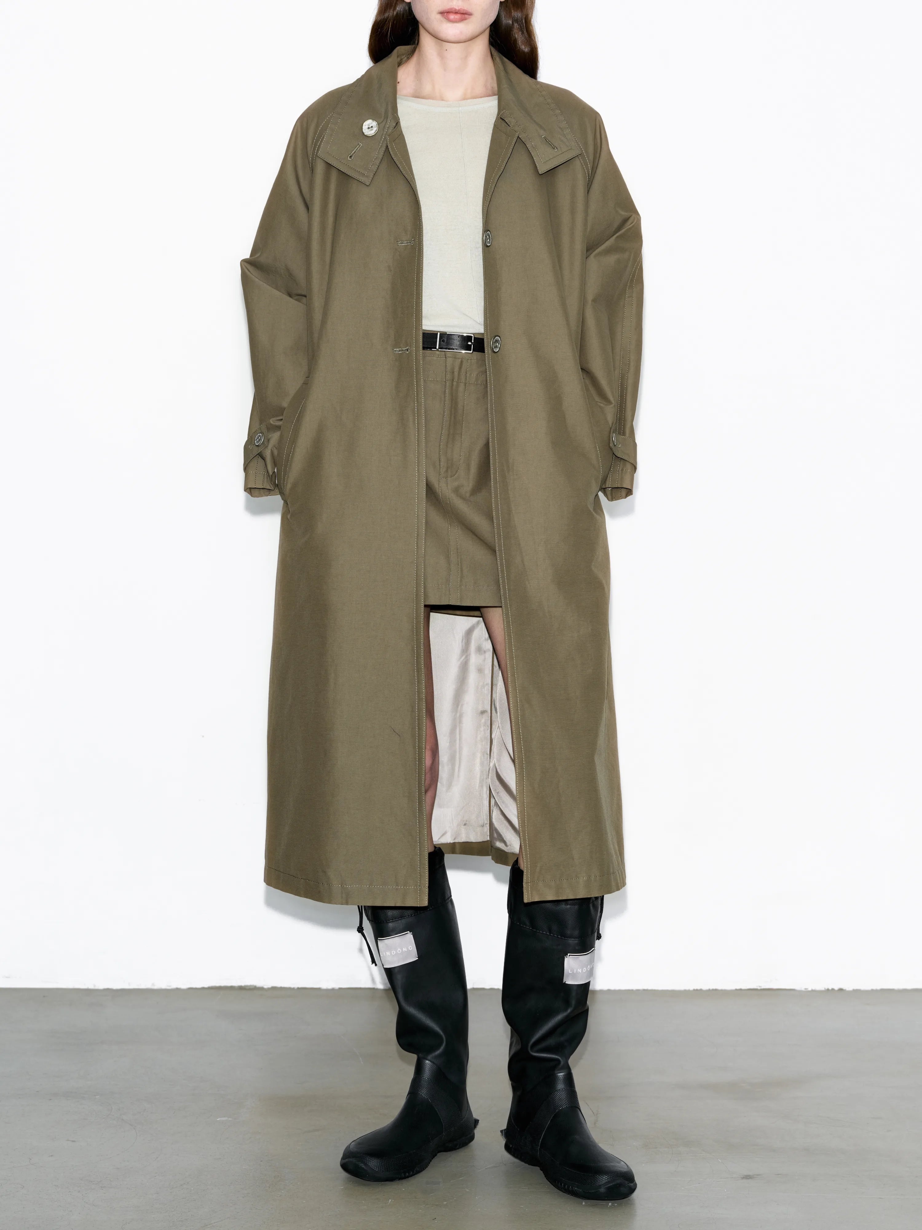 Model wearing army green trench coat, long wool-blend overcoat with layered back panels, oversized silhouette, styled with boots