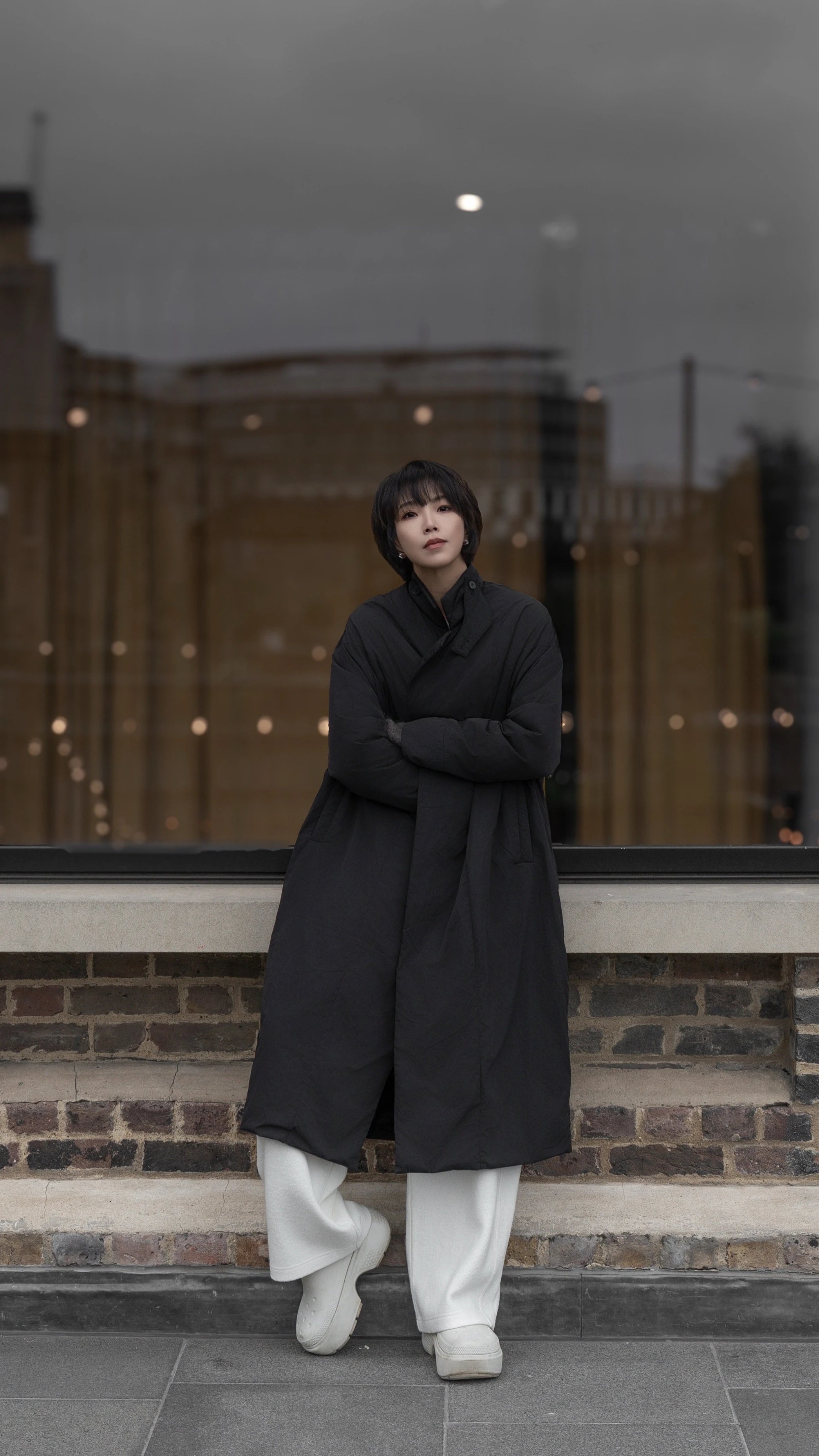  Model wearing a long black padded coat styled with white trousers, photographed in an urban street setting.