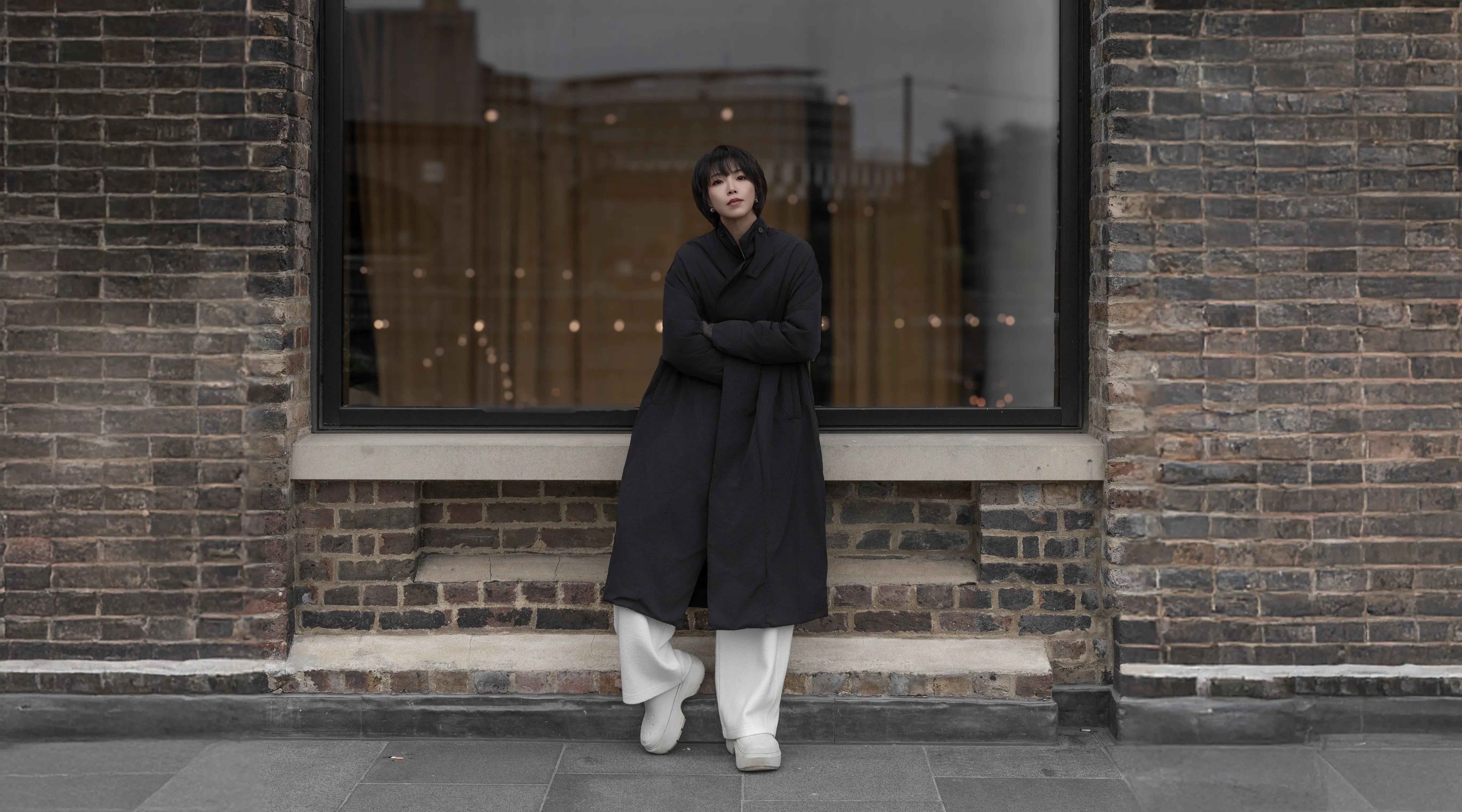 Model wearing a black oversized coat and white trousers standing against a brick wall background.