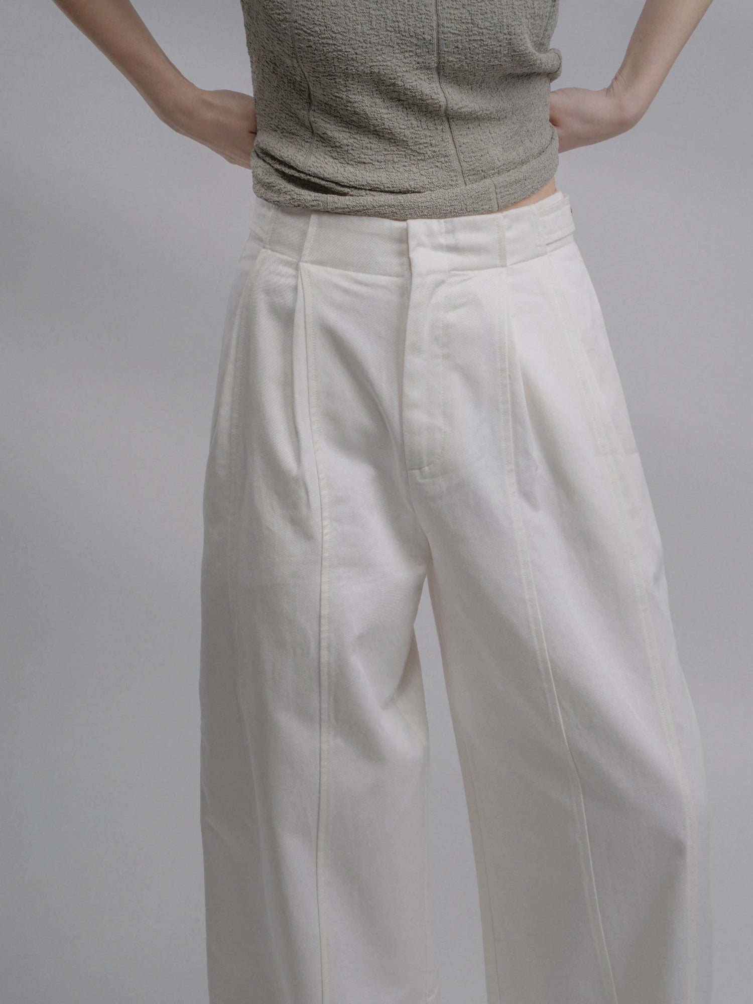 Close-up of waist and pleats on white loose-fit trousers, high-rise design with soft cotton texture and clean tailoring.