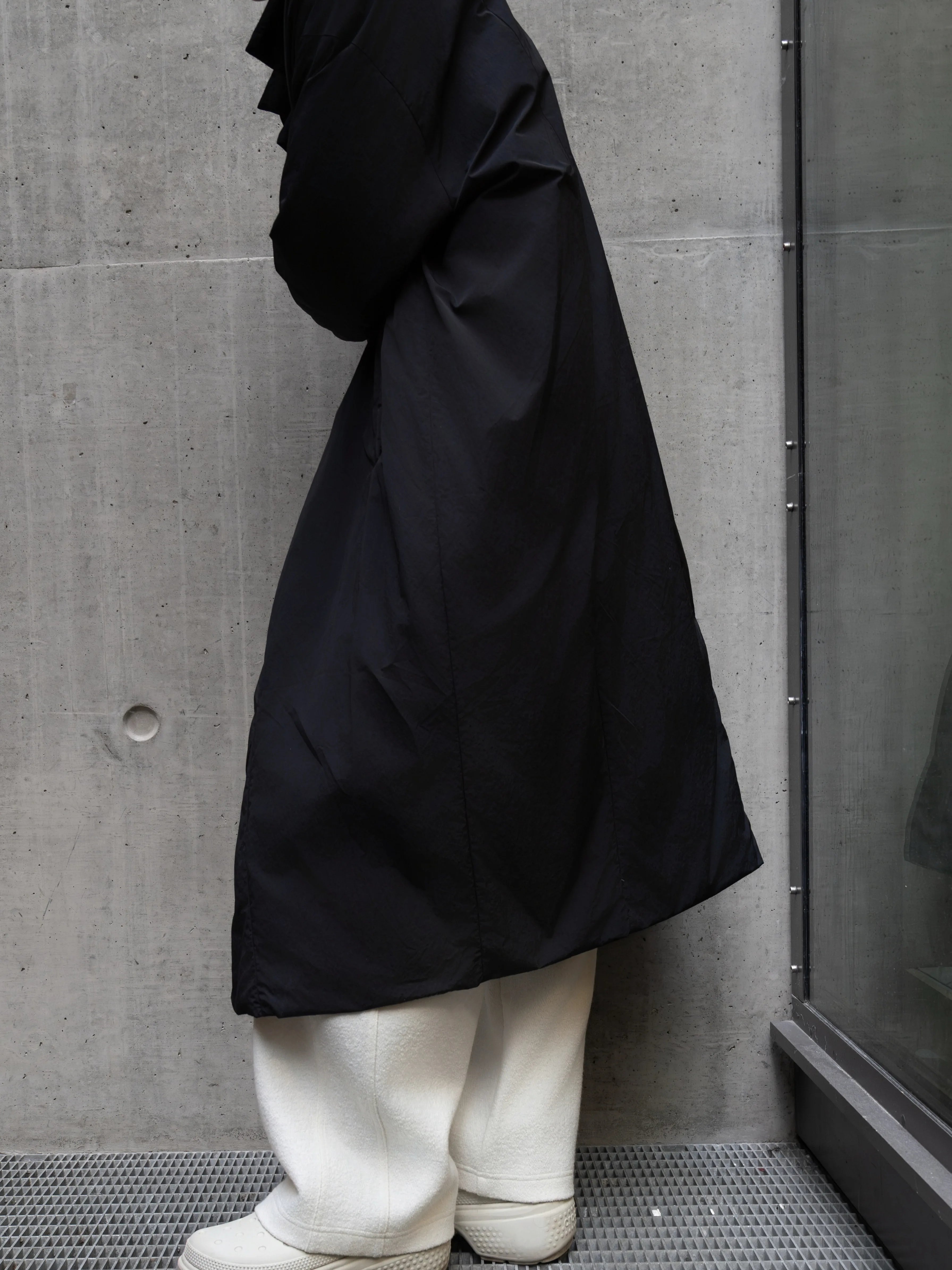 A side view of the black hooded down coat paired with white trousers, photographed against a concrete wall background.