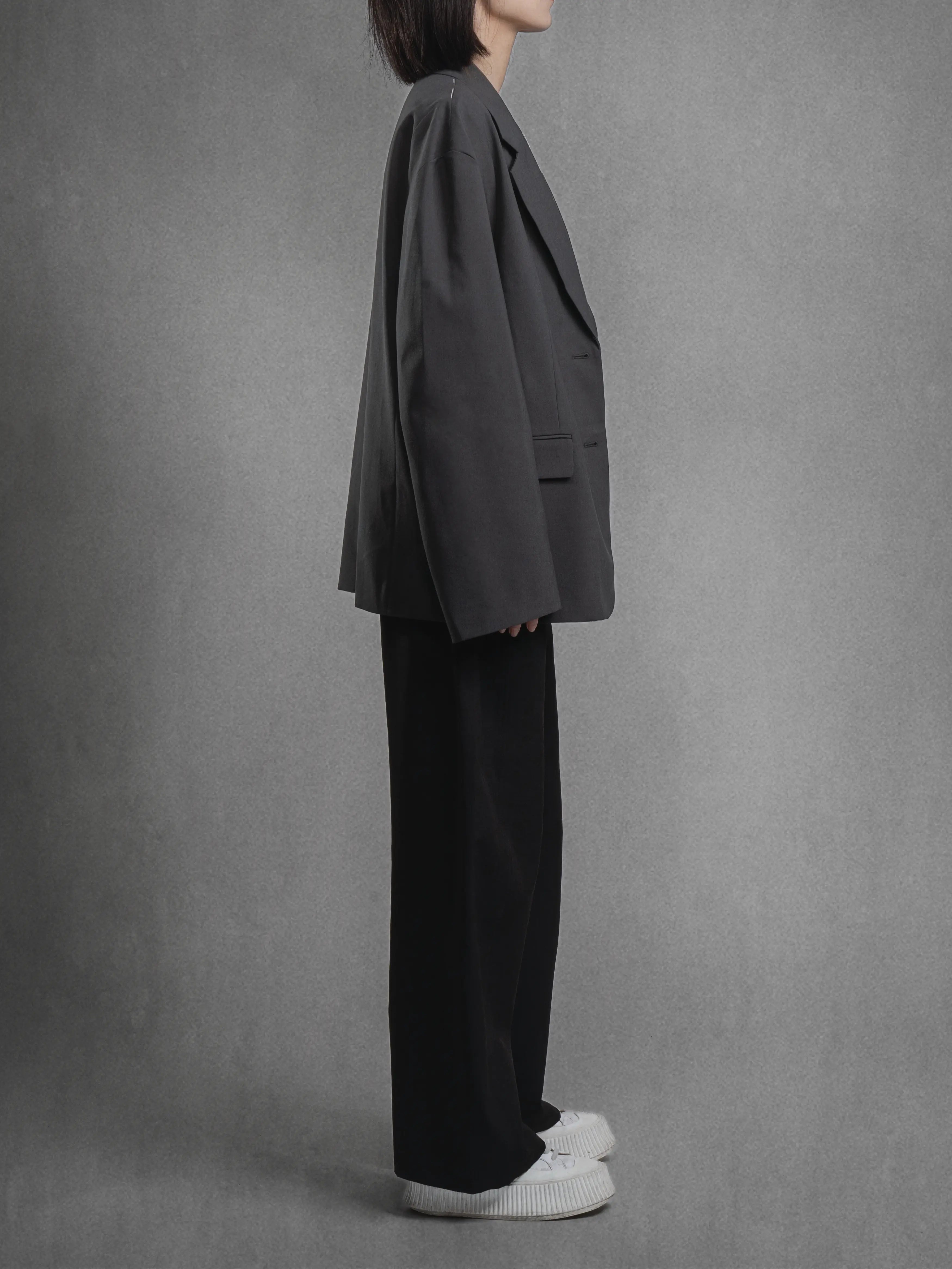 Side view of charcoal dropped shoulder blazer, soft-structured drape and exposed top-stitch detail.