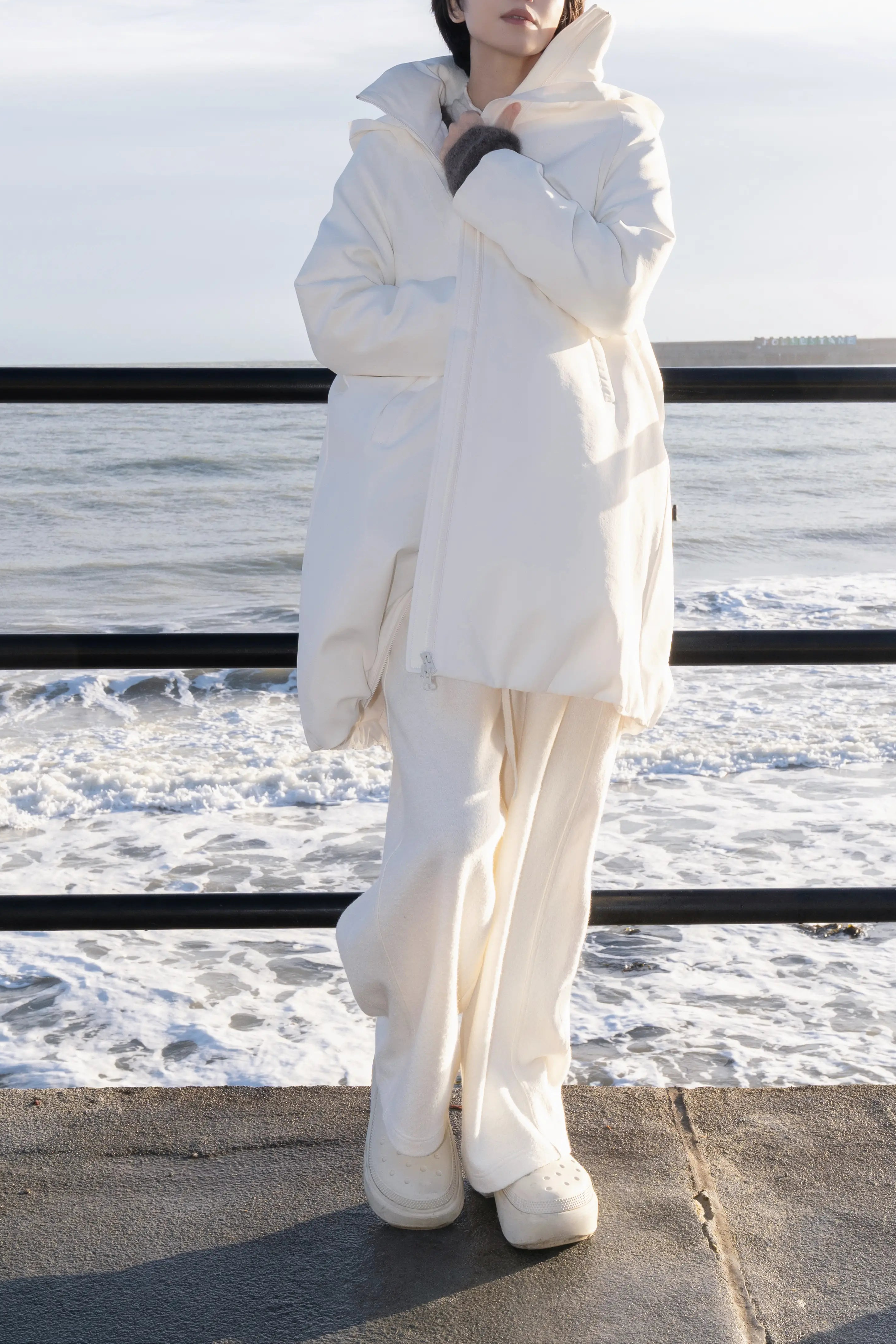 Model wearing an off-white hooded down coat standing by the seaside railings with waves in the background