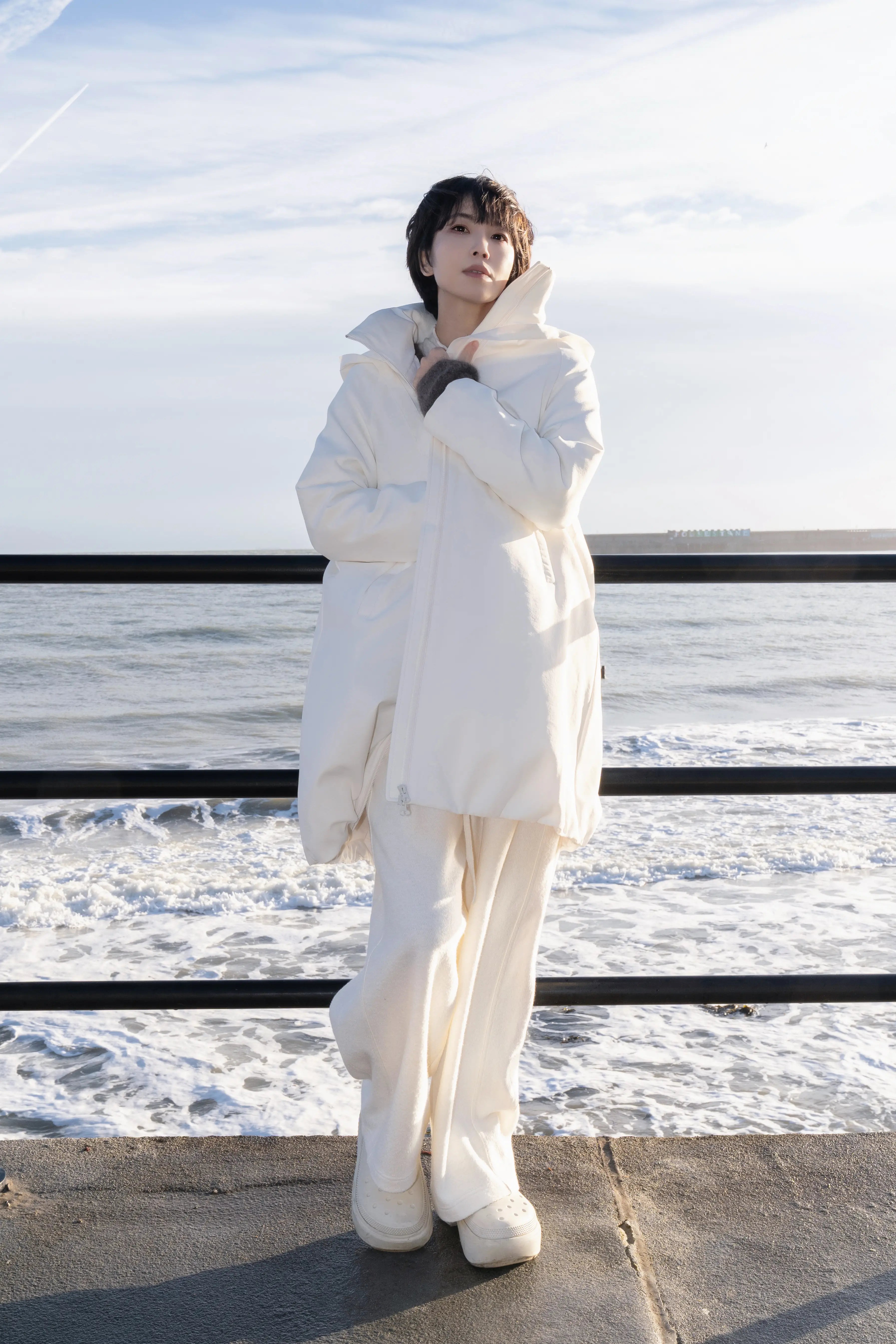 Model wearing an off-white hooded down coat standing by the seaside railings with waves in the background