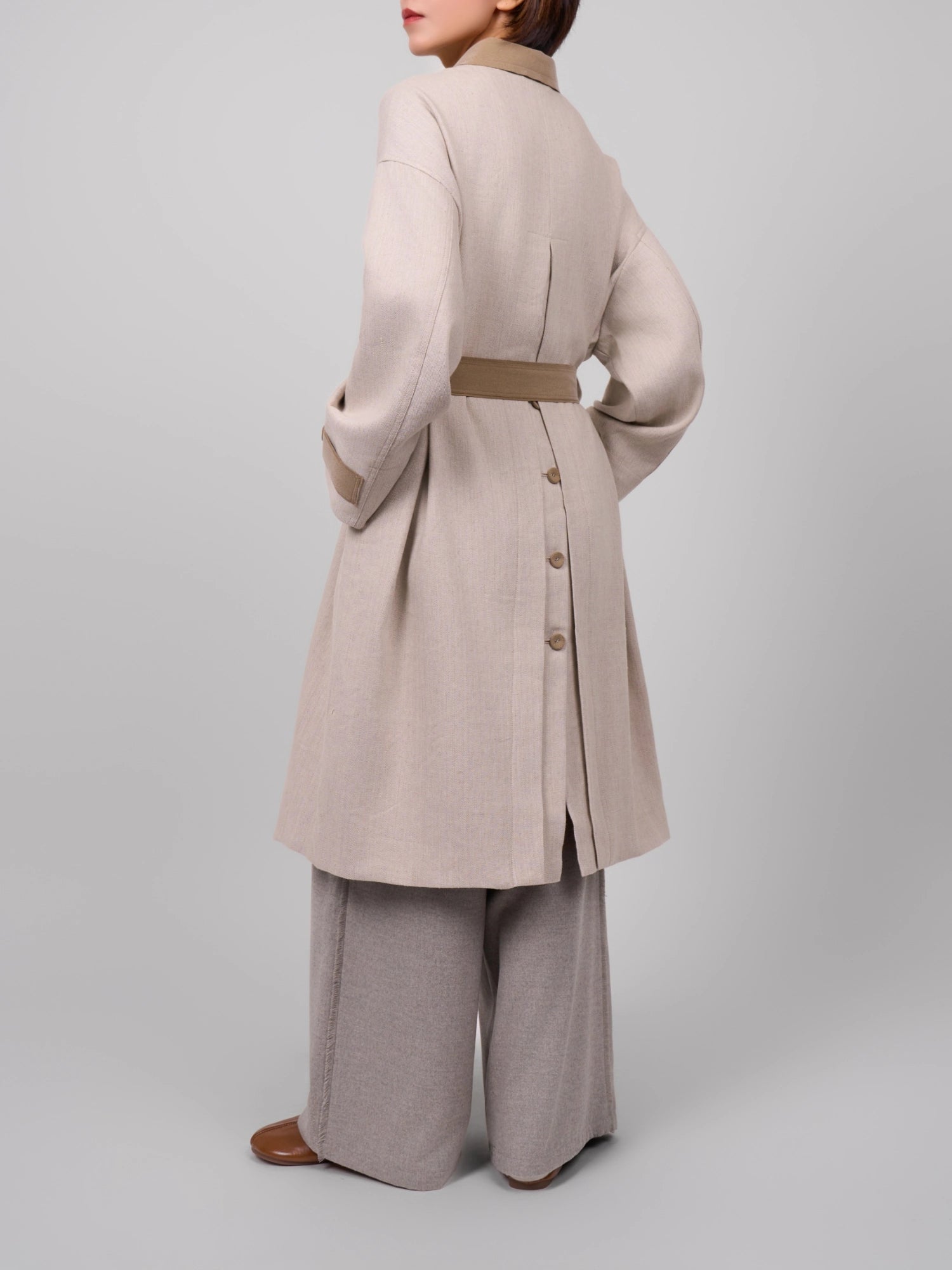 Back view of beige Rain Dew linen–wool coat, showing belt and buttoned back vent over wide-leg trousers.