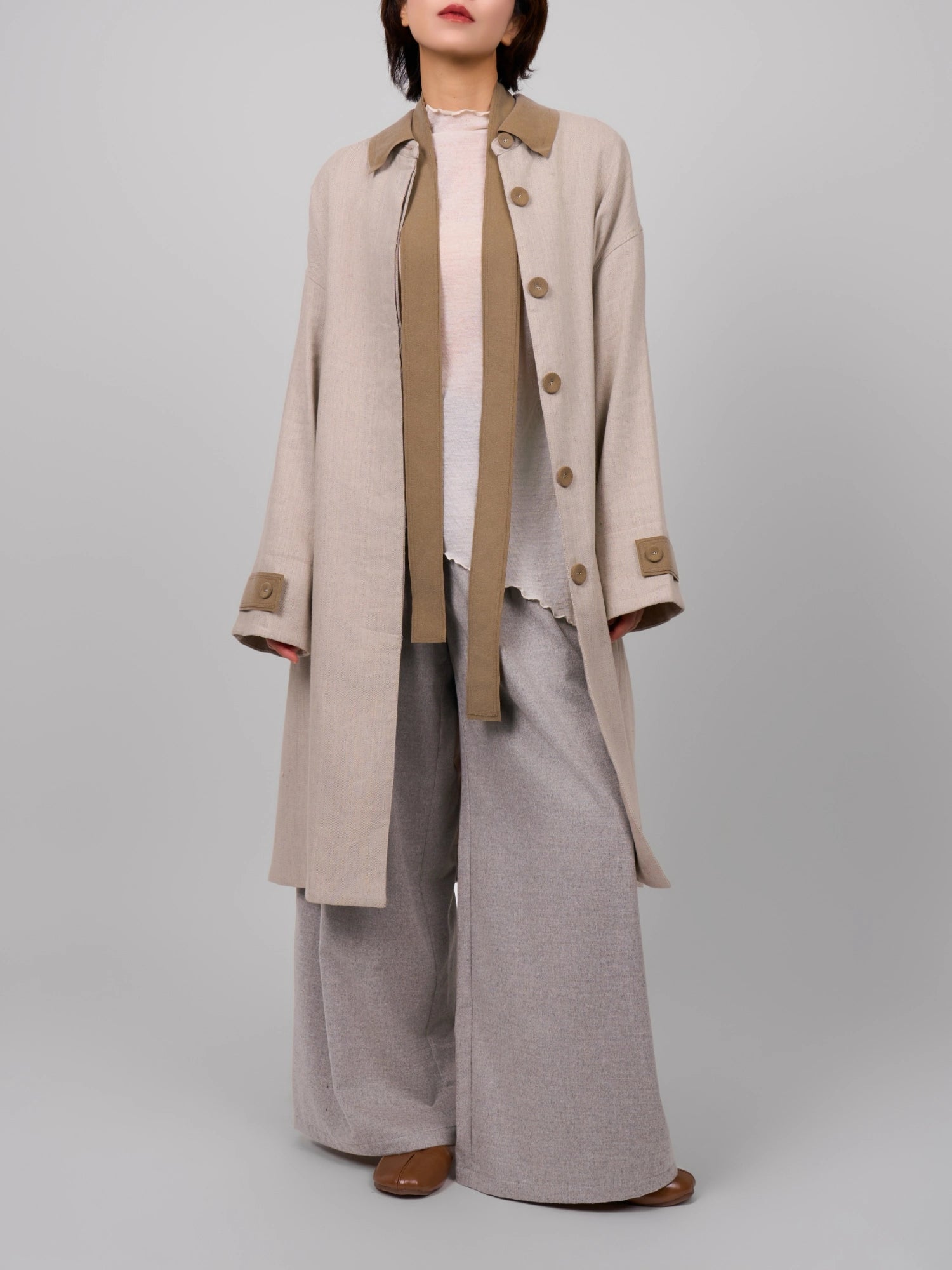 Model wearing beige Rain Dew linen–wool coat layered over knit and wide-leg trousers, full-body studio look.