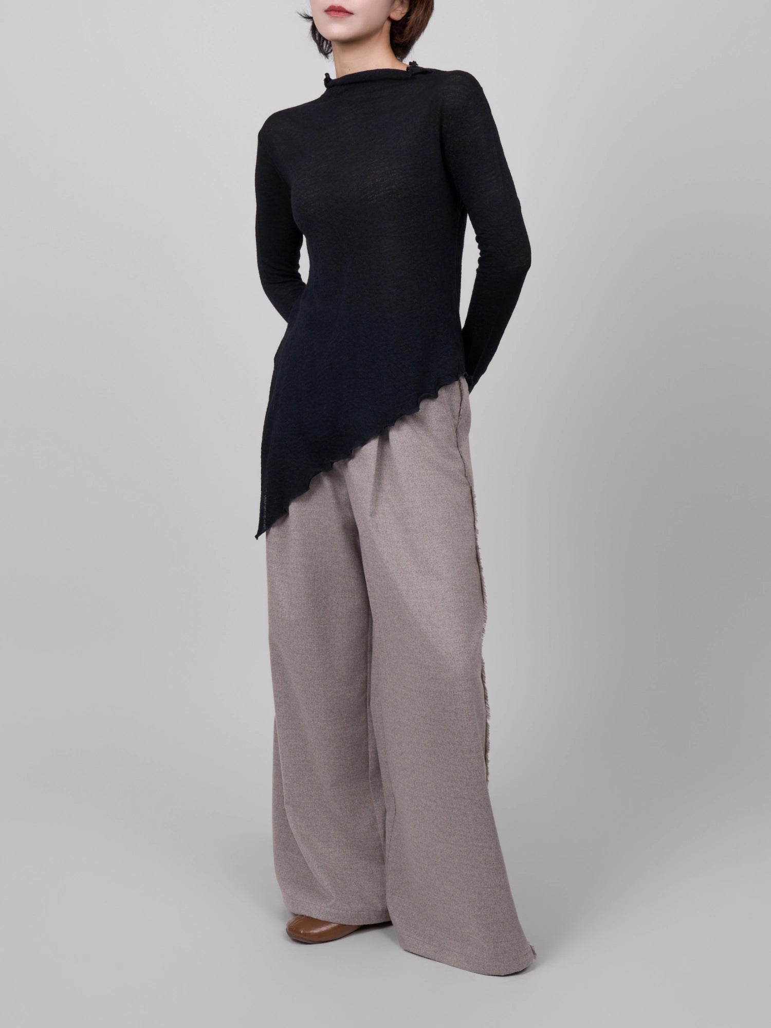 Model wearing black 100% wool base layer with asymmetric hem, full-body studio shot styled with wide-leg trousers.